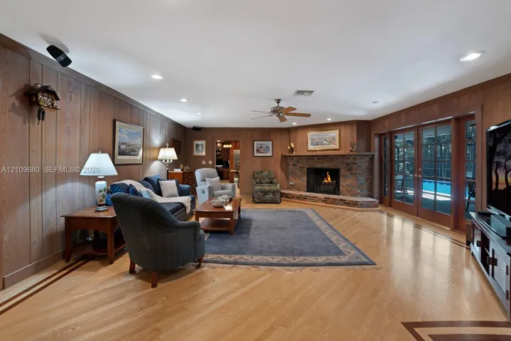 Living area featuring wood-paneled walls, wood flooring with inlaid border details, a corner stone fireplace with a raised brick hearth, a ceiling fan, and double glass-paneled doors