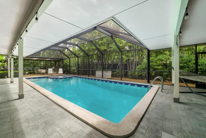 Screened-in pool area featuring an integrated spa, stone-tile decking, covered lanai, and surrounding lush greenery