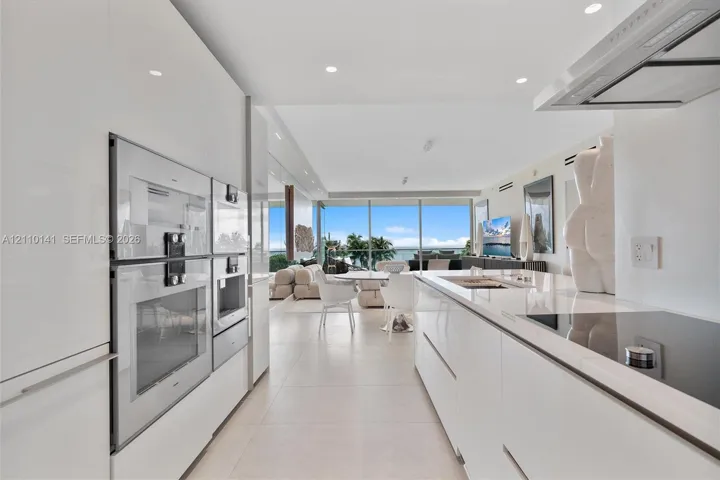 Contemporary kitchen featuring white high-gloss cabinetry, stainless steel built-in double wall ovens, a seamless solid-surface island countertop, integrated cooktop, and floor-to-ceiling glass paneling
