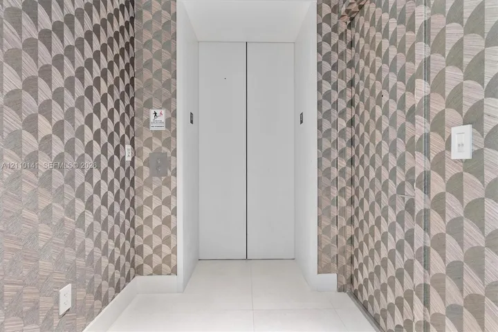Private elevator entry featuring white double doors, geometric patterned wall covering, and neutral tile flooring