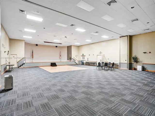 Expansive multi-purpose hall featuring a raised stage, inset hardwood floor segment, recessed lighting, and patterned modular carpeting