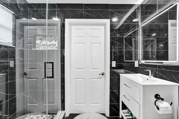 Bathroom featuring black, veined-stone wall tiling, glass-enclosed shower with a built-in recessed niche, white vanity with a rectangular sink, and an illuminated mirror