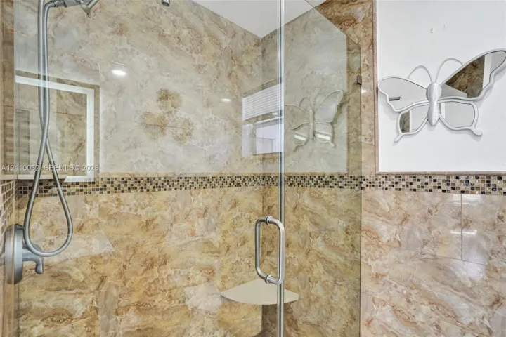 Walk-in shower featuring floor-to-ceiling stone-finish tile, glass enclosure, handheld shower fixture, built-in corner shelving, and decorative mosaic accent tile inlay