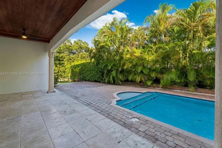 Covered patio with natural stone flooring and wood-paneled ceiling, featuring a swimming pool, brick paver decking, and perimeter landscaping