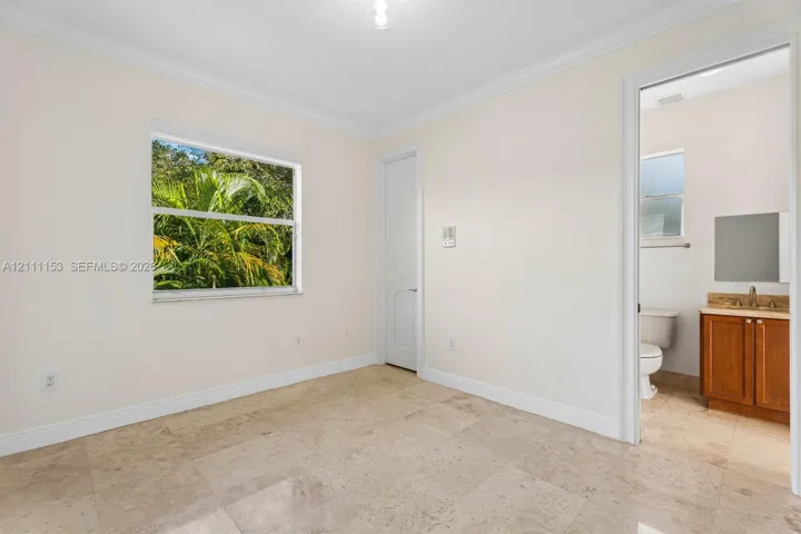 Interior space featuring stone tile flooring, crown molding, a window with exterior greenery, and a doorway revealing a vanity with wood-finish cabinetry