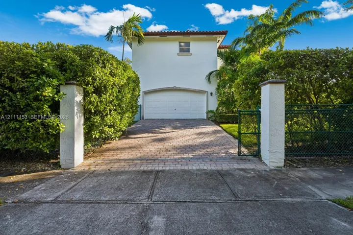 Stucco exterior featuring a paver driveway, arched garage entry, decorative stucco pillars, and mature landscaping