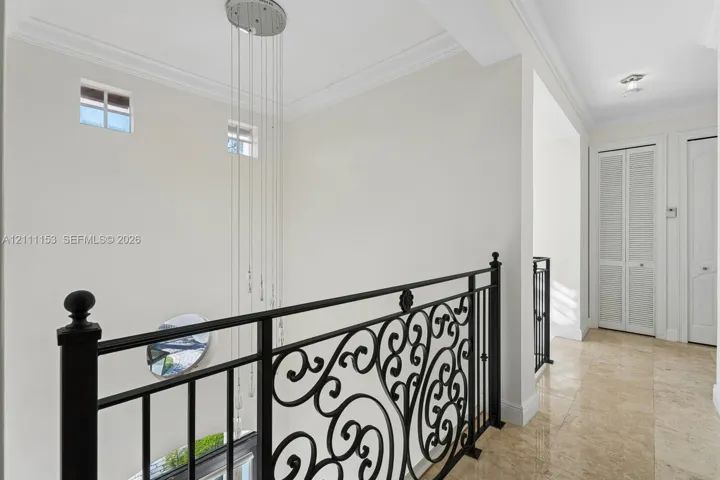 Hallway featuring polished stone flooring, intricate black iron railing, multi-tiered pendant lighting, crown molding, and louvered closet doors
