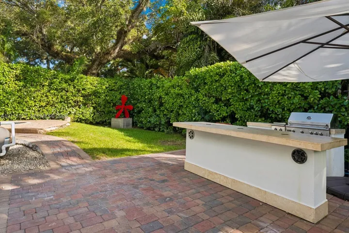 Outdoor kitchen featuring a built-in grill and stone-capped bar, set upon a red brick paver patio, surrounded by mature privacy hedging and a manicured lawn
