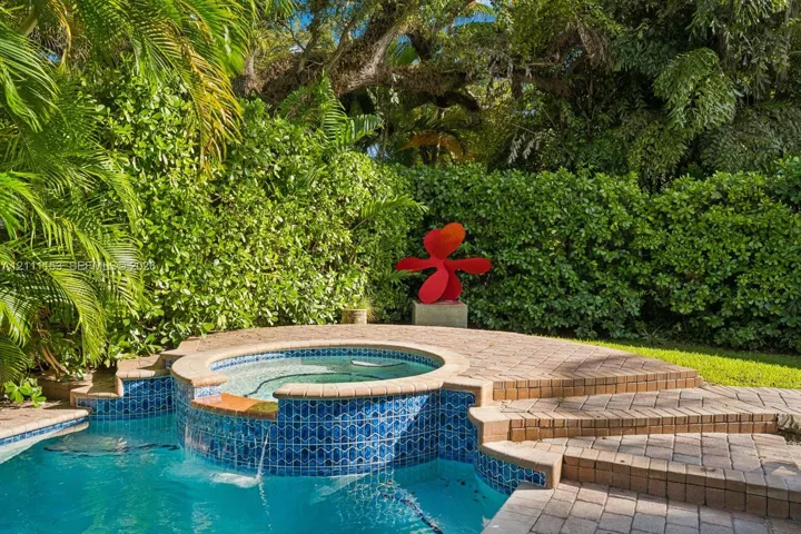 Swimming pool featuring a raised, tiled spa with a water spillover, surrounded by a tiered brick-paver patio and lush perimeter hedges