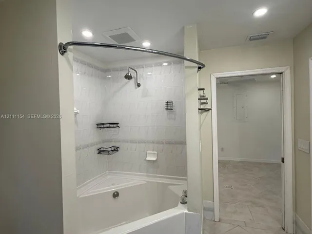 Corner soaking tub with integrated shower head, white tiled surround with decorative inlay, curved shower rod, and recessed ceiling lighting