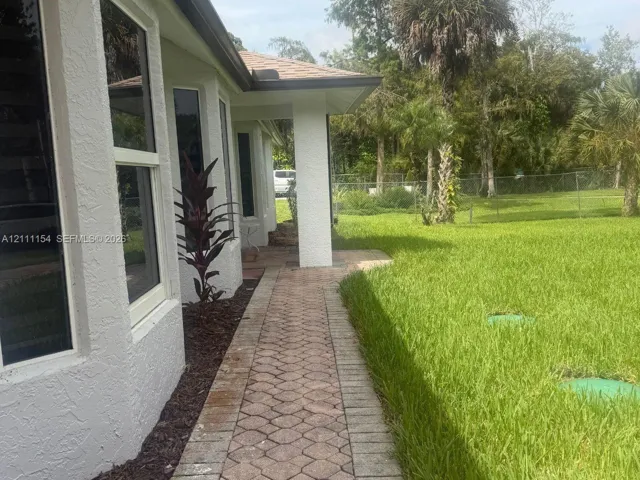 Stucco exterior with a paved stone walkway, overhanging roofline, and expansive lawn with perimeter fencing
