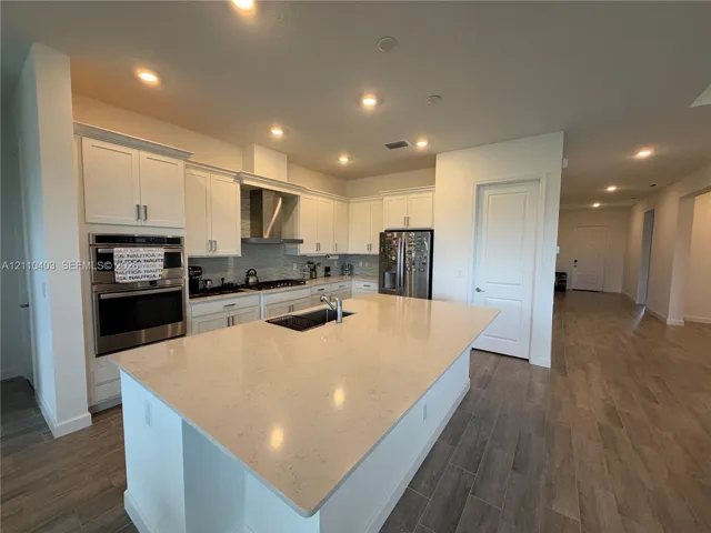 Kitchen featuring a spacious central island with a quartz countertop, white shaker-style cabinetry, stainless steel double wall ovens, built-in stainless steel refrigerator, and wood-finish tile flooring