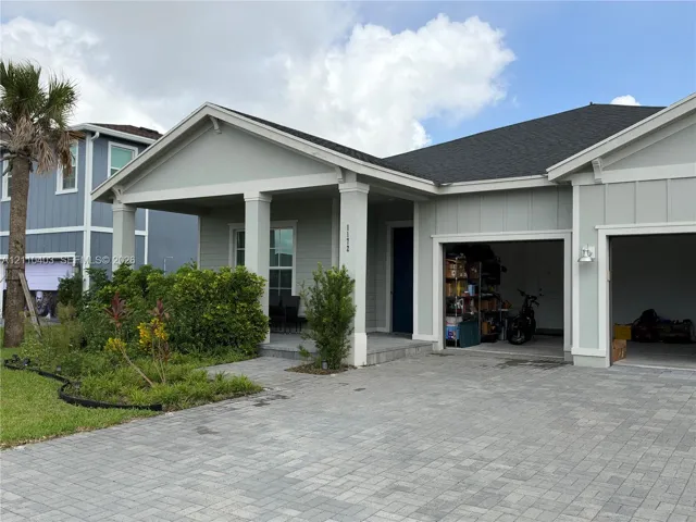 Single-story exterior featuring light-colored siding, a covered front porch with square columns, a multi-car garage, and a paved stone driveway