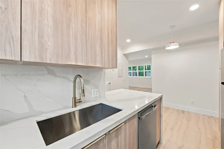 Kitchen featuring light wood-finish cabinetry, stone-surface countertops, full-height stone-surface backsplash, undermount stainless steel sink with a high-arc faucet, and recessed lighting