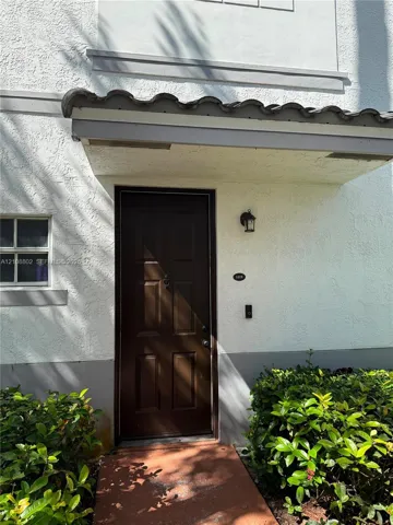 Stucco exterior featuring a dark brown multi-panel entry door, arched tile awning, integrated wall-mounted exterior lighting, and a paved walkway