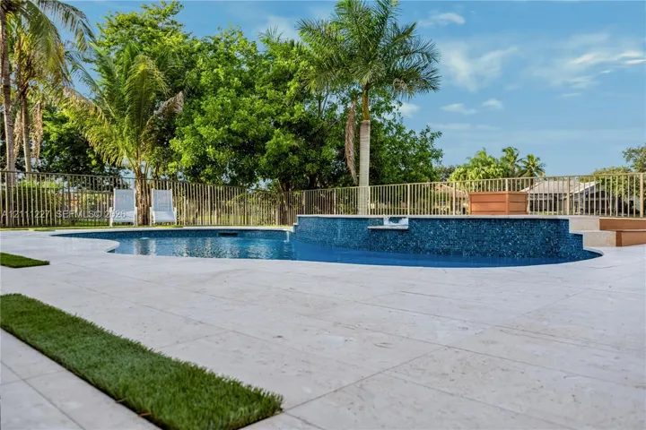 Custom pool featuring a raised tiled spillway, light-toned stone paver decking, integrated grass inlay accents, and a metal perimeter fence