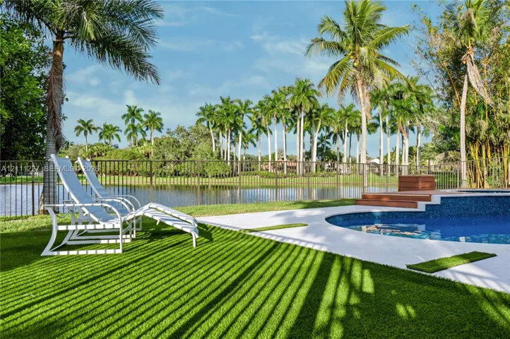 Outdoor pool area featuring a blue-tiled swimming pool, synthetic turf lawn, metal perimeter fencing, and mature palm trees