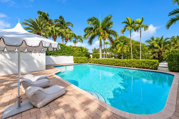 In-ground swimming pool featuring a brick-paver patio, perimeter privacy hedging, and mature palm trees