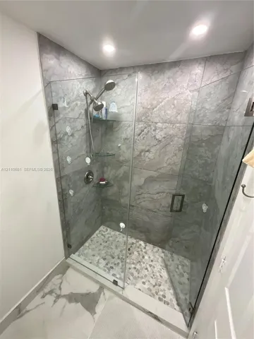 Walk-in shower featuring large-format stone-finish wall tile, pebble-tile flooring, frameless glass enclosure, and built-in glass corner shelving