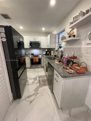 Kitchen featuring white shaker-style cabinetry, stone-finish countertops, stainless steel appliances, stacked-tile backsplash, and large-format porcelain tile flooring