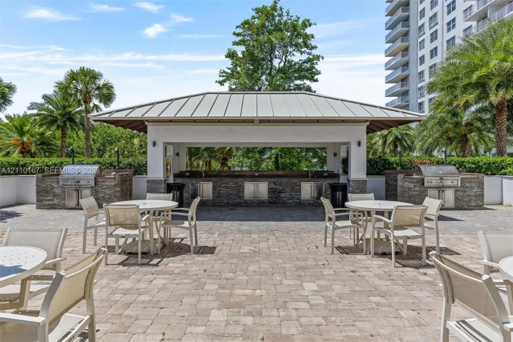 Outdoor pavilion featuring a stone-clad kitchen island, dual built-in stainless steel grills, a metal-seam roof, and extensive stone paver patio space
