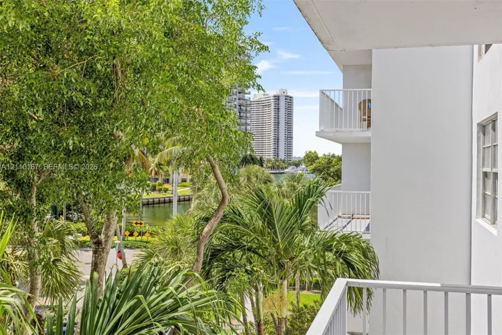 Private balcony with white metal railings, overlooking lush landscaping, mature trees, and a canal