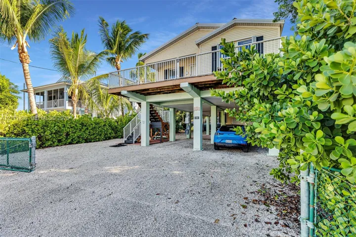 Elevated coastal-style residence featuring horizontal siding, a covered ground-level parking area, a spacious second-story balcony with metal railings, and a gravel driveway