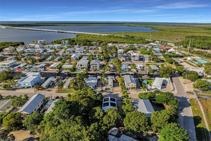 Aerial perspective of coastal residential property featuring mature trees, a rooftop solar panel system, paved street access, and proximity to expansive waterfront and causeway