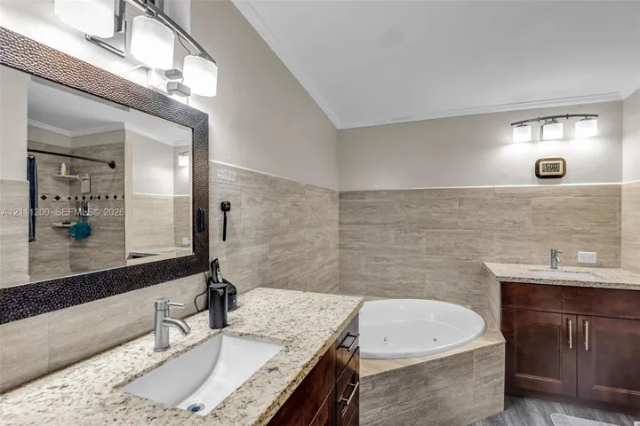 Bathroom featuring a jetted soaking tub with stone tile surround, dual dark wood vanities, stone countertops with integrated sinks, and polished chrome fixtures