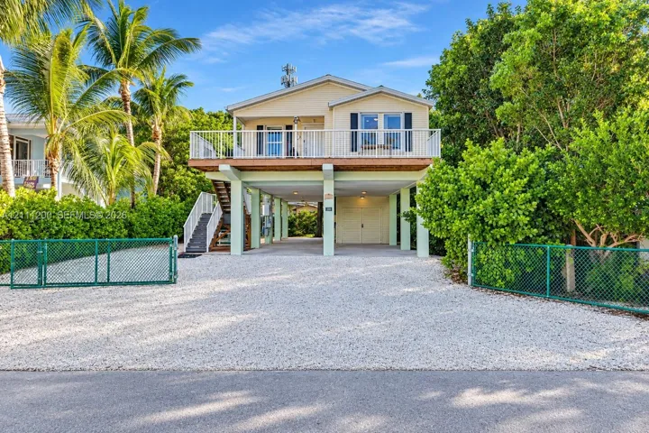 Elevated residence featuring a raised deck with railing, gravel driveway, external staircase, and chain-link perimeter fencing