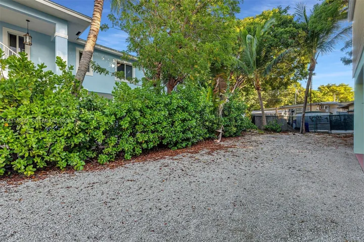 Side exterior area featuring a gravel surface, mature palm trees, and established shrubbery surrounding the light blue siding of the home