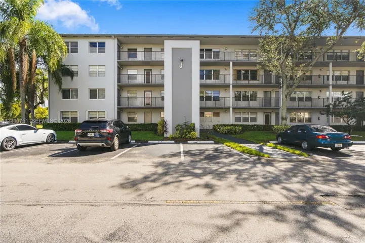 Multi-story residential building exterior featuring concrete construction, metal balcony railings, assigned surface parking, and mature landscaping