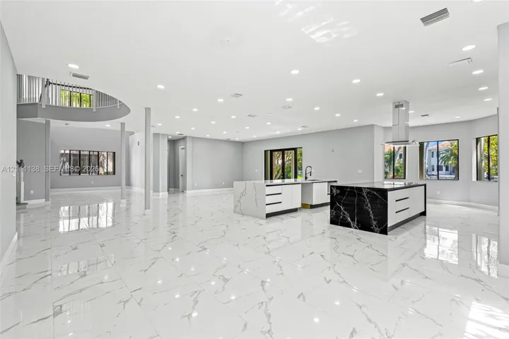 Open-concept living space featuring large-format tile flooring, dual kitchen islands with contrasting waterfall-edge stone surfaces, recessed ceiling lighting, and a curved staircase with a metal balustrade