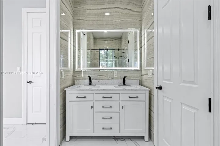 Double vanity featuring a white cabinetry base, stone-look countertop, integrated dual sinks, and matte black widespread faucets, accented by full-height linear pattern tile walls and a modern LED-backlit mirror
