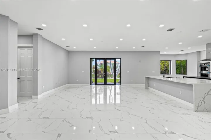Open-concept interior featuring large-format polished stone flooring, recessed ceiling lighting, a waterfall-edge kitchen island, and double glass-paned doors