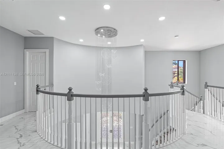 Curved architectural wall featuring a multi-tiered crystal chandelier, polished stone flooring, and a balustrade staircase with wood-finish handrails