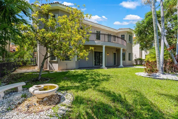 Two-story stucco exterior featuring a curved upper-level balcony with metal railing, classic columns, stone-accented landscaping, and a tiered fountain
