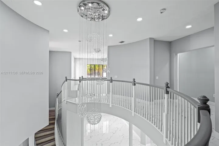 Curved balcony featuring a multi-tiered crystal chandelier, white stone-finish flooring with grey veining, and a white balustrade with a contrasting handrail