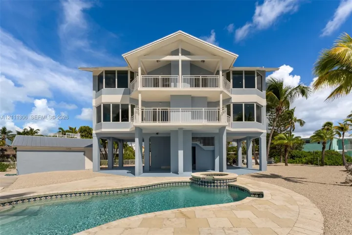 Three-story stilt structure featuring multi-level covered balconies, an inground swimming pool with integrated spa, stone-paver pool deck, and a detached garage