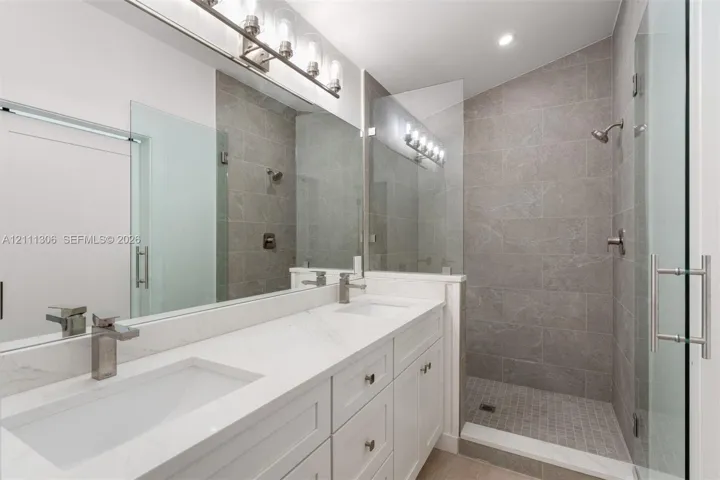 Dual-sink vanity with stone-finish countertops and chrome fixtures, paired with a walk-in shower featuring large-format tile surround, mosaic floor tiling, and a glass enclosure