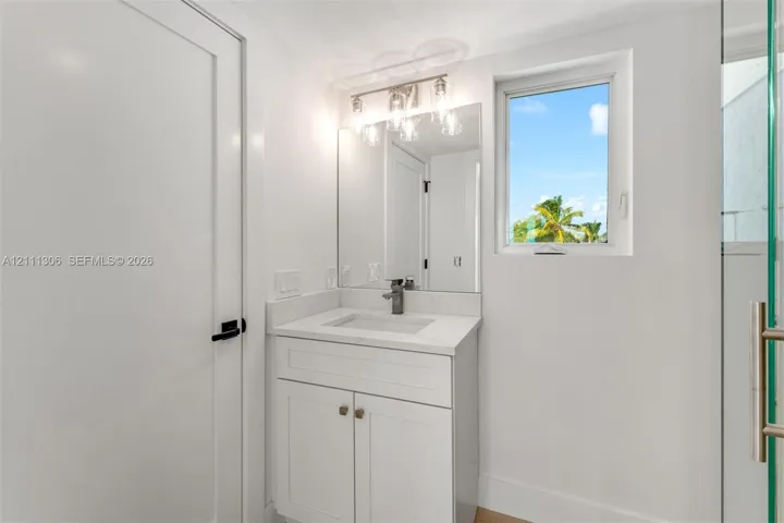 White shaker-style vanity with a solid surface countertop, integrated rectangular sink, three-light fixture with glass shades, wall-mounted mirror, and glass-enclosed shower