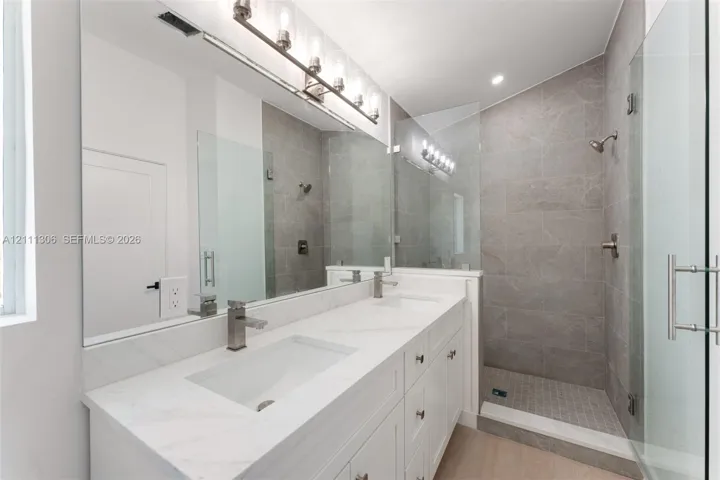 Double-vanity with stone countertop, walk-in shower featuring tile surround and glass enclosure, large wall-mounted mirror, and multi-bulb vanity lighting