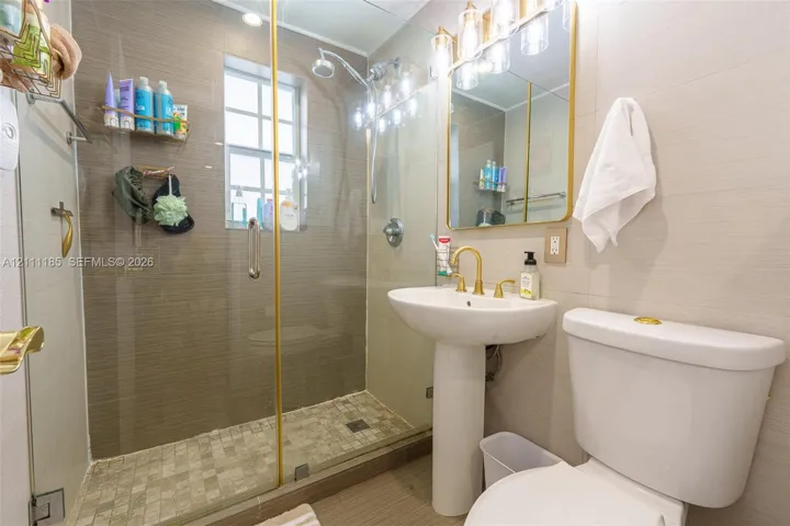 Full bathroom featuring a walk-in shower with a glass enclosure, gold-tone hardware, pedestal sink, and decorative wall-mounted lighting