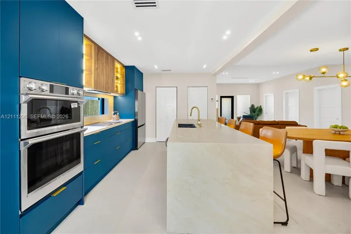 Kitchen featuring custom blue cabinetry, wood-finish shelving with integrated lighting, a waterfall-edge stone island, built-in stainless steel double oven, and recessed ceiling lighting