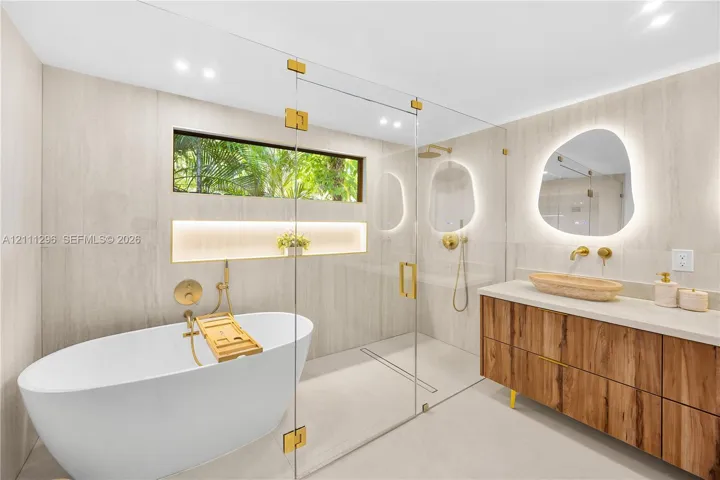 Freestanding soaking tub with gold-tone fixtures, walk-in glass-enclosed shower, wall-to-wall stone-style tile, wood-finish floating vanity with vessel sink, and organic-shaped backlit mirrors