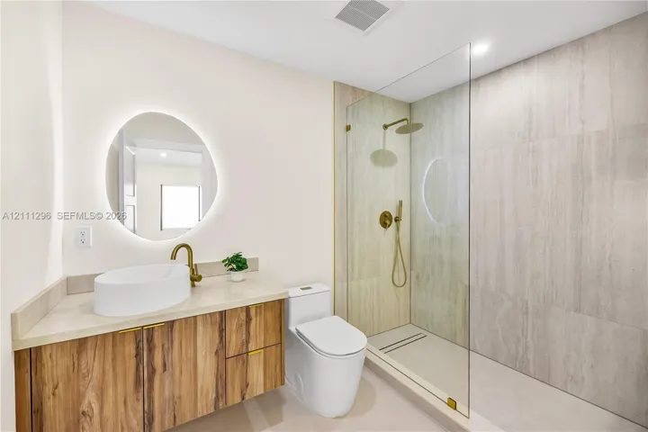 Floating wood-finish vanity with an oval vessel sink, back-lit circular mirror, walk-in shower with floor-to-ceiling tile, and brass-finish fixtures