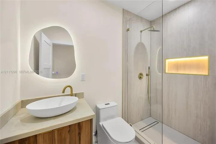 Bathroom featuring an asymmetrical backlit mirror, wood-finish vanity with a vessel sink, brass fixtures, glass-enclosed shower with tiled walls, and a recessed shower niche with integrated lighting