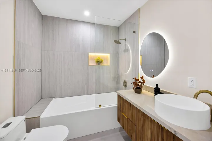 Bathroom featuring a wood-finish vanity with a vessel sink, quartz-style countertop, a walk-in shower with a glass panel and integrated recessed niche, and a circular LED-backlit mirror