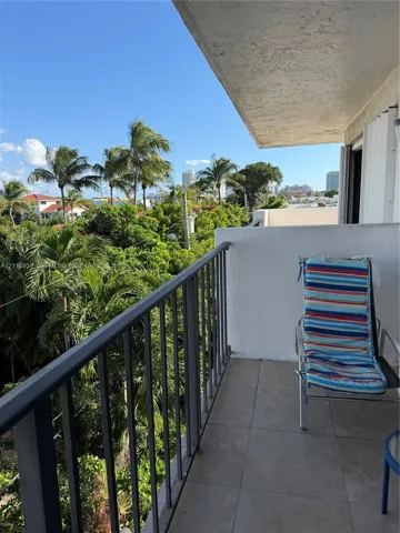 Covered balcony with a dark metal railing, tiled flooring, and high-reaching exterior walls, complemented by mature palm trees and lush greenery