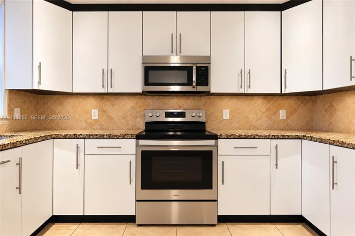White shaker-style cabinetry with modern hardware, stone-surface countertops, neutral diagonal-set tile backsplash, and stainless steel appliances including a built-in microwave and range